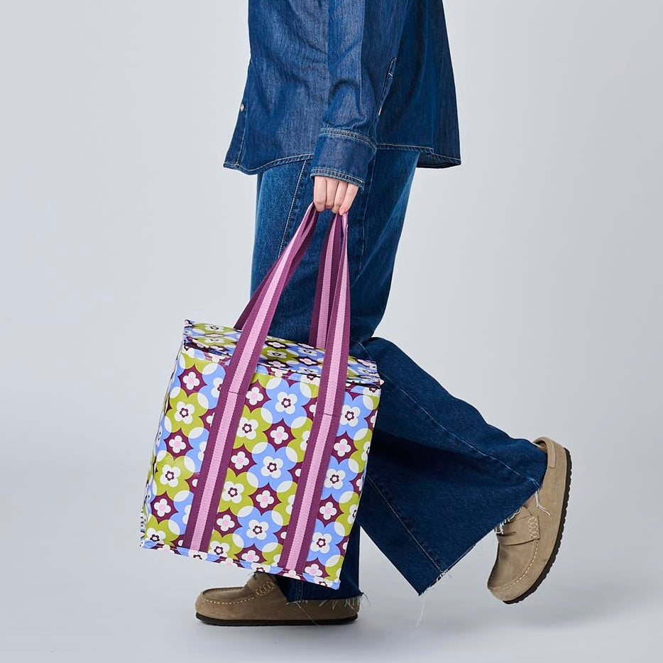 A woman in a blue denim jacket and wide-leg jeans walks in profile while carrying a colorful patterned tote bag with purple handles against a plain light gray studio background.