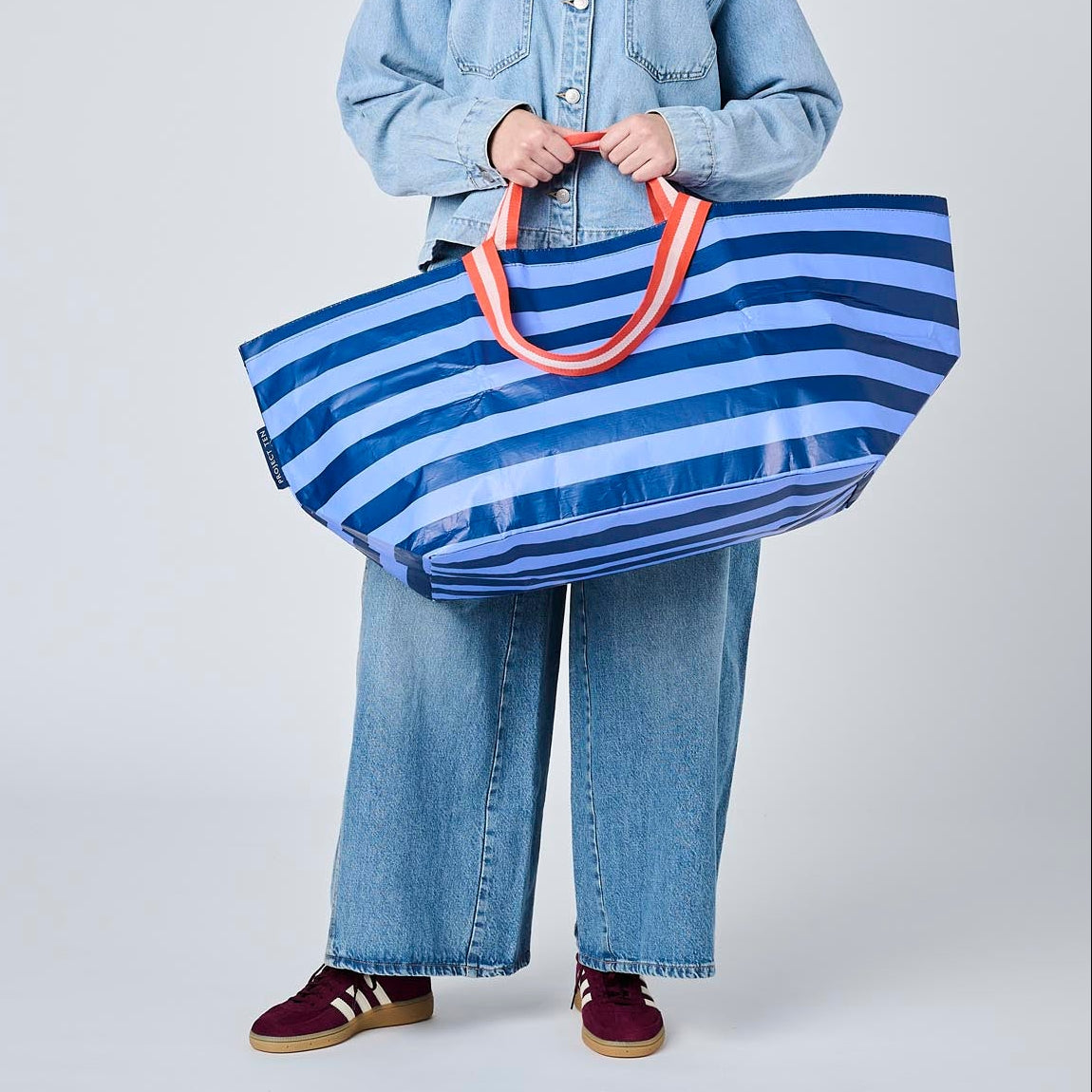 A model wearing a light blue denim jacket and wide-leg jeans holds a large blue and white striped tote bag with orange handles against a light gray studio background.