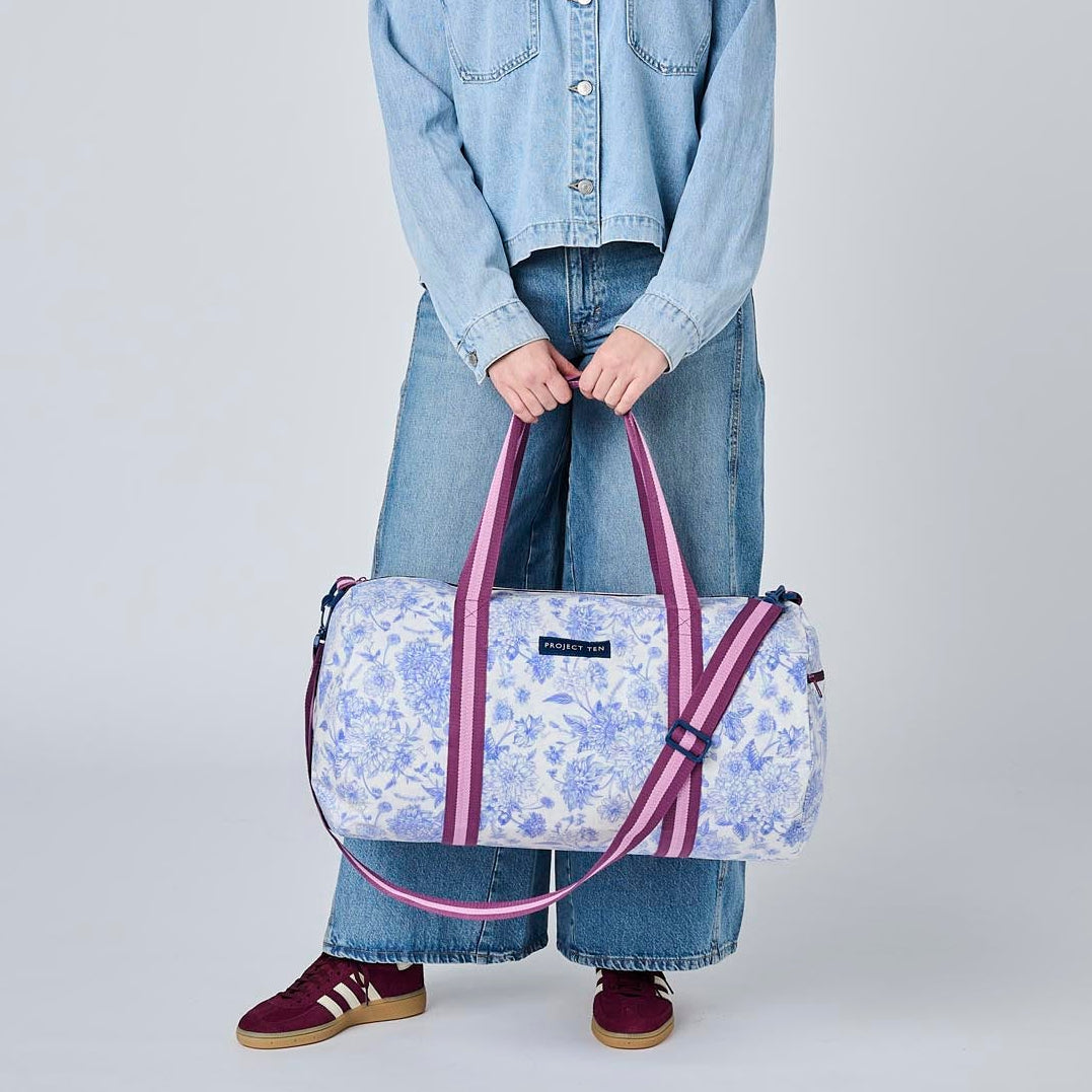 A model stands against a light gray studio background wearing a light blue denim jacket, wide-leg jeans, and burgundy sneakers, while holding a light blue and white floral print duffel bag with pink handles.