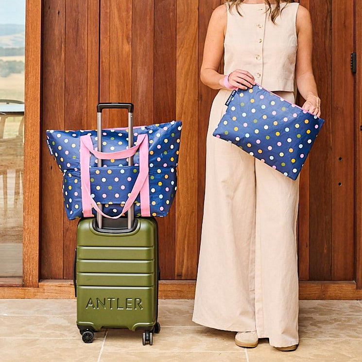 Woman standing with a green suitcase and two polka dot bags in front of a wooden building.
Bag on suitcase is zip medium tote and lady is holding the large zip pouch.