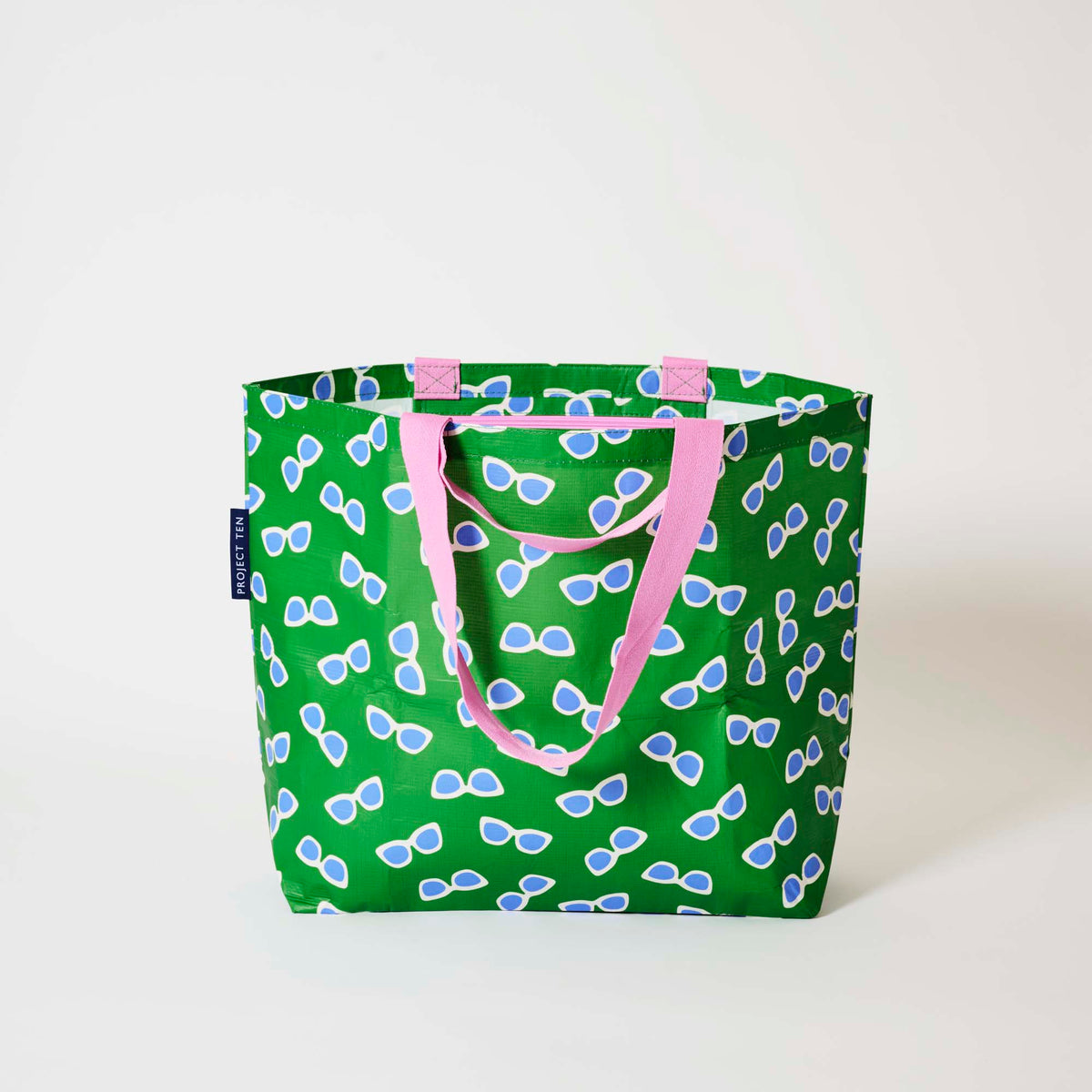 Our Sunglasses Medium Tote in green with a scattered print of ecru framed sunglasses with blue lenses, and pink straps, shown open against a plain background.