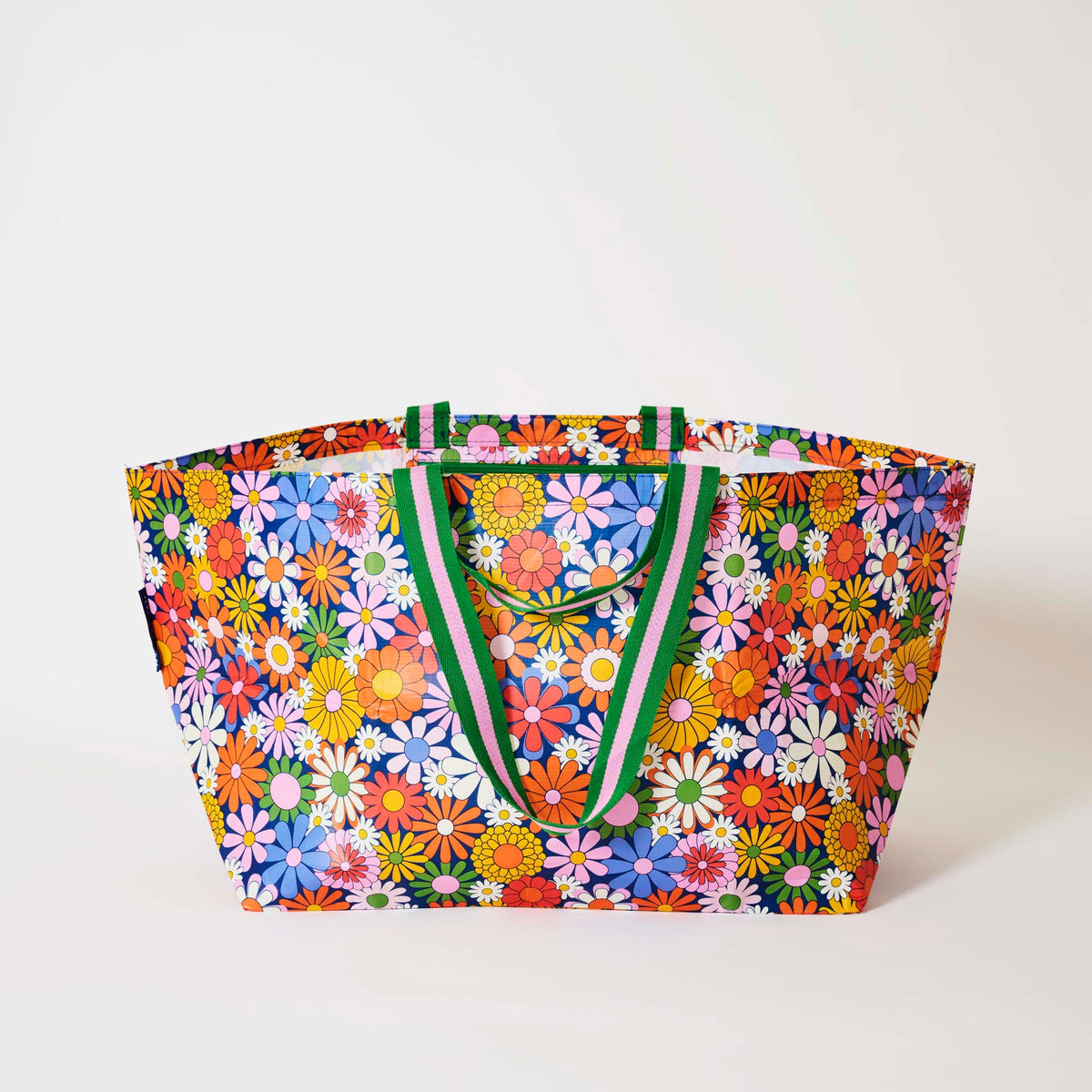 Our Coastal Floral Oversized Tote covered in bright retro daisies in orange, pink, yellow and blue on a navy background, with green and pink striped straps, shown open against a plain background.