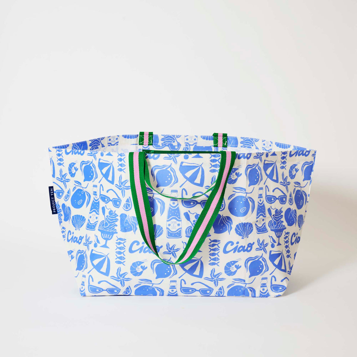 Our Feast Oversized Tote in a blue and ecru print of lemons, seafood, gelato, soda bottles, sunglasses, beach umbrellas and the word "Ciao", with green and pink striped straps, shown open against a plain background.