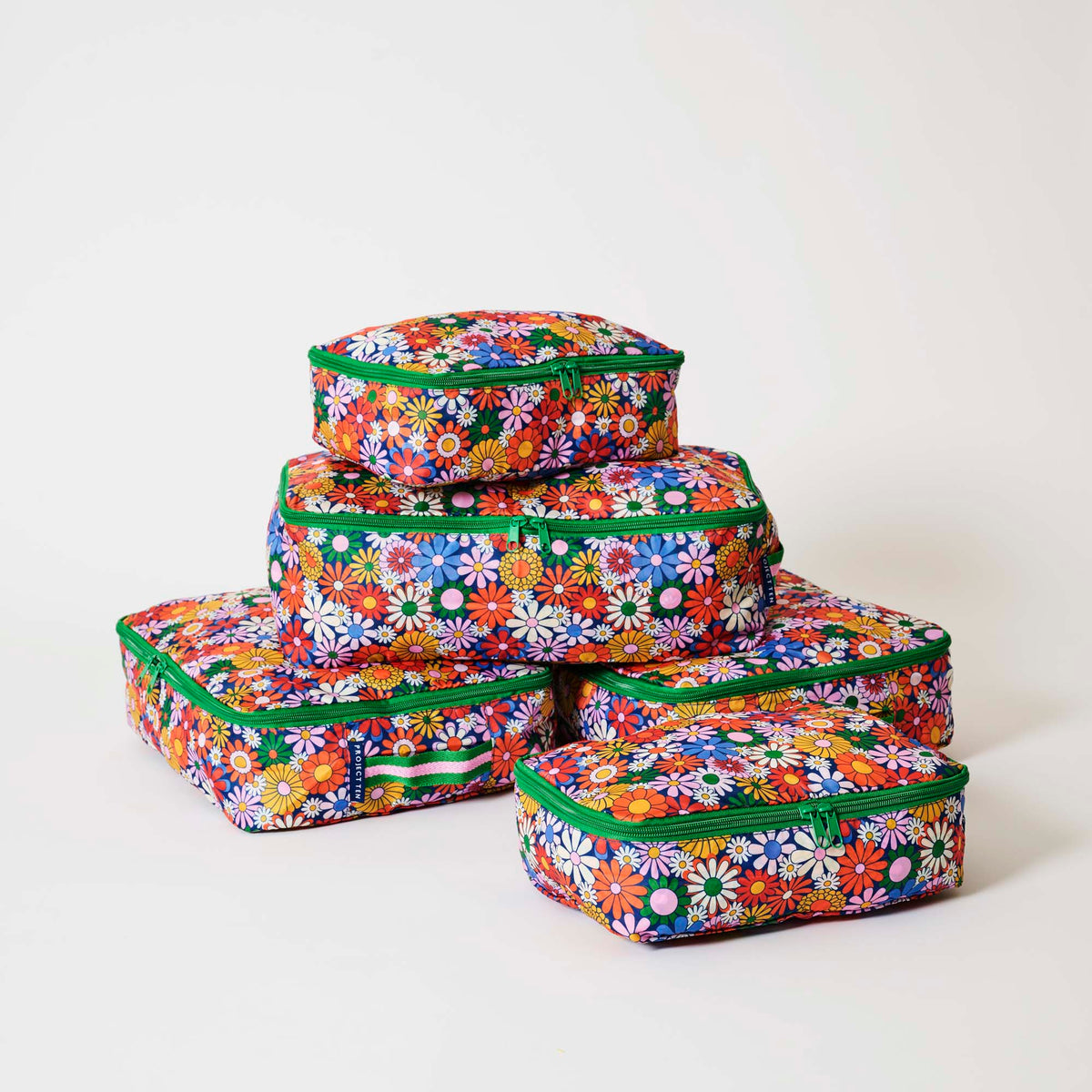 Our Coastal Floral Packing Cubes, a set of five zip-up packing cubes in different sizes, in a retro daisy print of orange, red, pink, yellow, blue and white flowers on a navy background, with green zips and green and pink striped carry handles.
