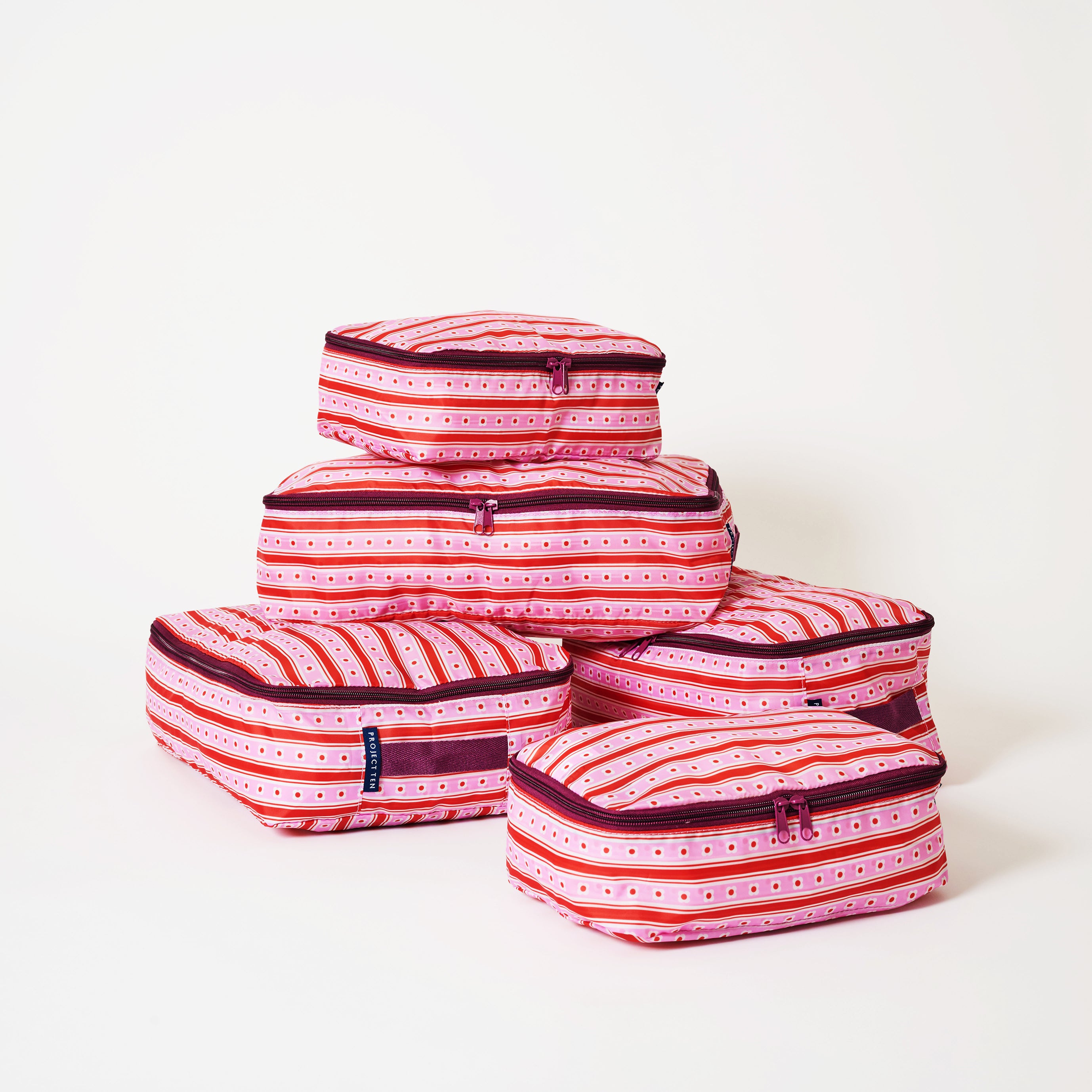 A set of five packing cubes stacked in a pyramid arrangement, featuring a vibrant pink, red, and white striped pattern with small dotted details and dark purple trim.
- Project Ten