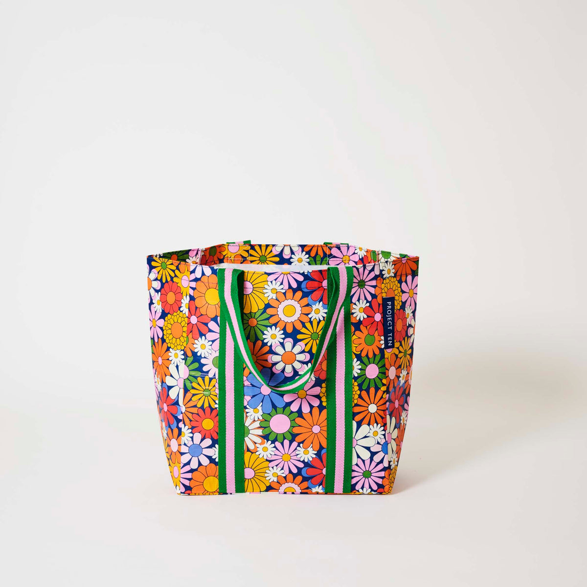 Our Coastal Floral Shopper Tote covered in bright retro daisies in orange, pink, yellow and blue on a navy background, with green and pink striped straps that run down the front and back, shown open against a plain background.