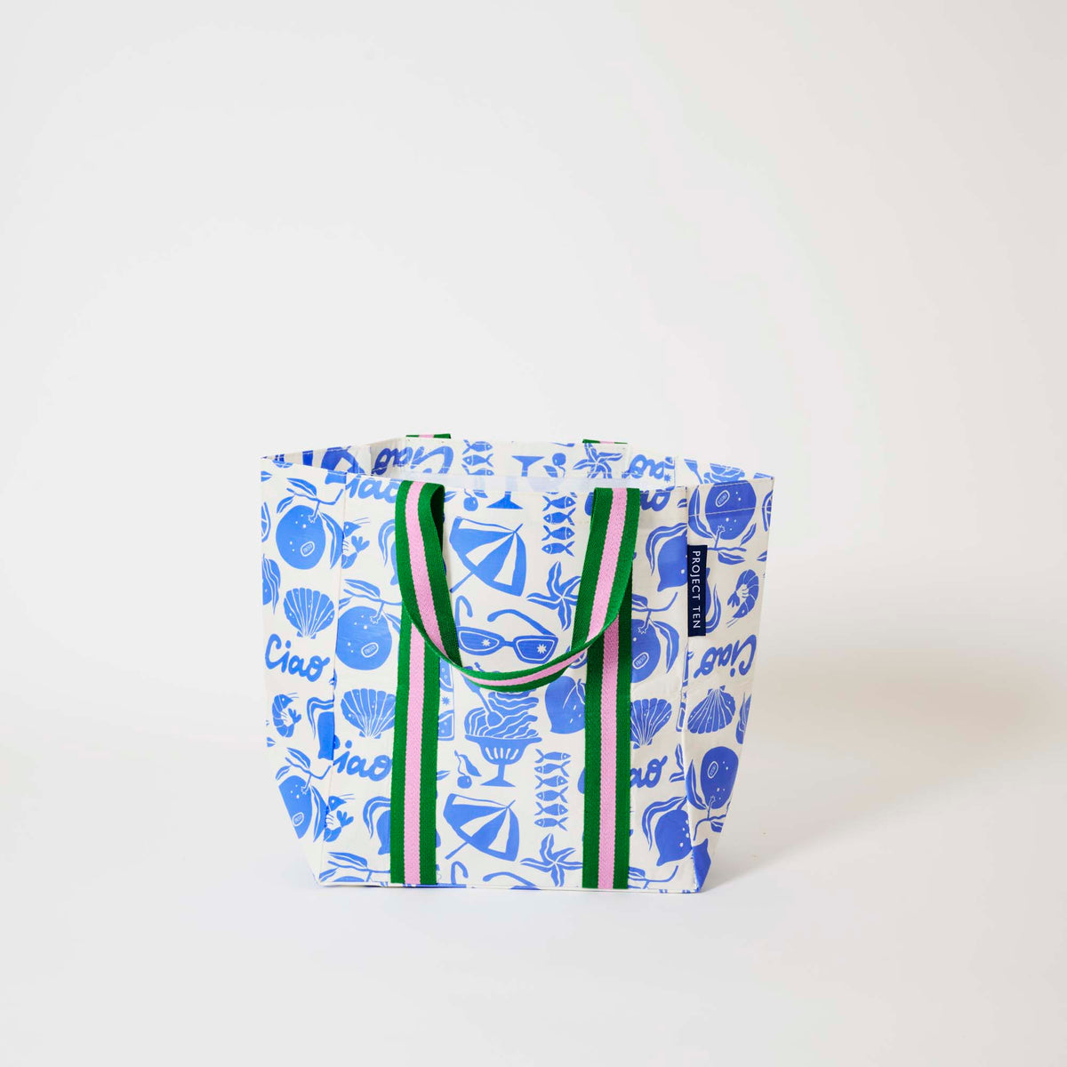 Our Feast Shopper Tote in a blue and ecru print of lemons, seafood, gelato, sunglasses, beach umbrellas and the word "Ciao", with green and pink striped straps that run down the front and back, shown open against a plain background.