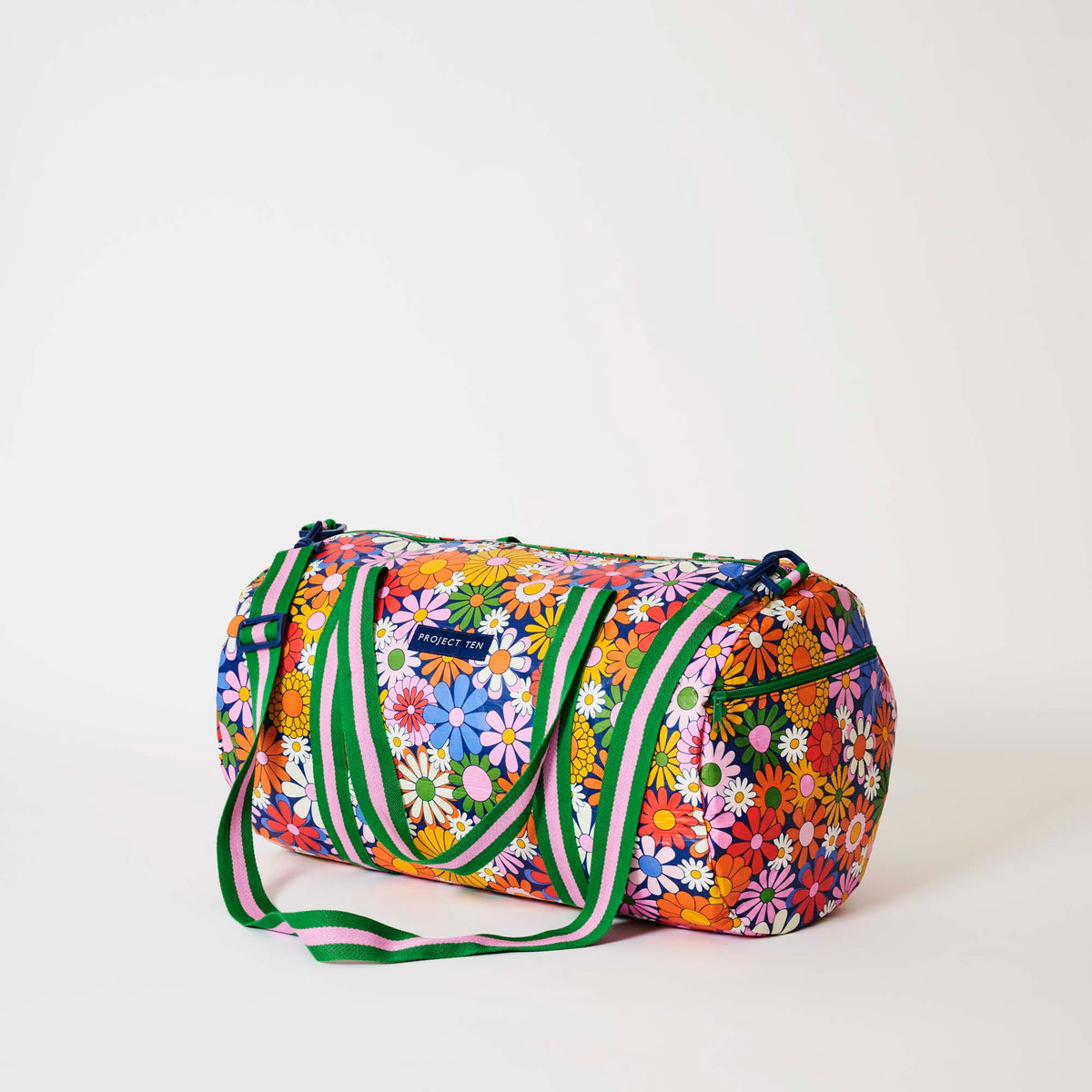 Our Coastal Floral Barrel Bag, a cylindrical duffle-style bag in a retro daisy print of orange, red, pink, yellow, blue and white flowers on a navy background, with two green and pink striped carry handles, a detachable matching shoulder strap and a green zip.