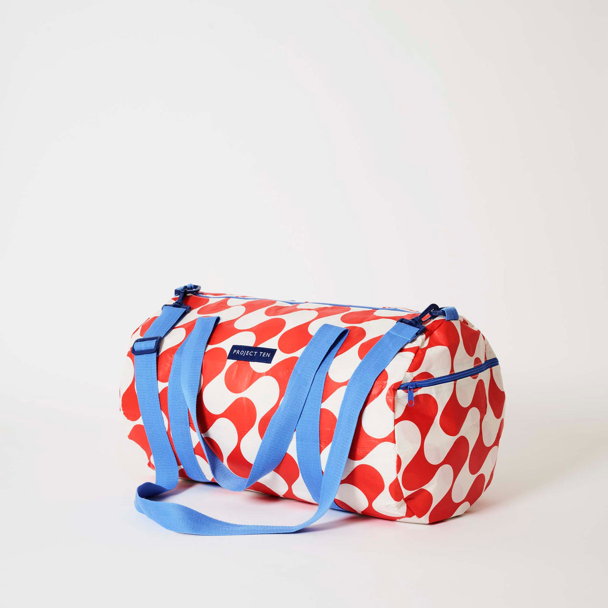 Our Red Ripple Barrel Bag, a cylindrical duffle-style bag in a bold geometric wave print of red and ecru, with two blue carry handles, a detachable blue shoulder strap and a blue zip.