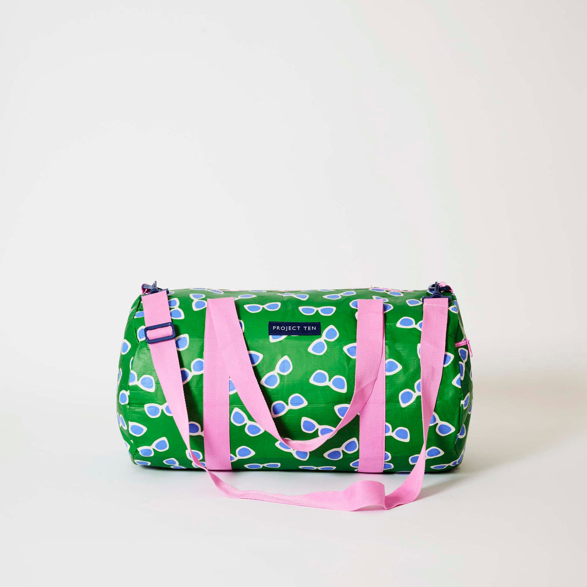 Our Sunglasses Barrel Bag, a cylindrical duffle-style bag in green with a scattered print of blue-lensed, white-framed sunglasses, two pink carry handles, a detachable pink shoulder strap and a pink zip.