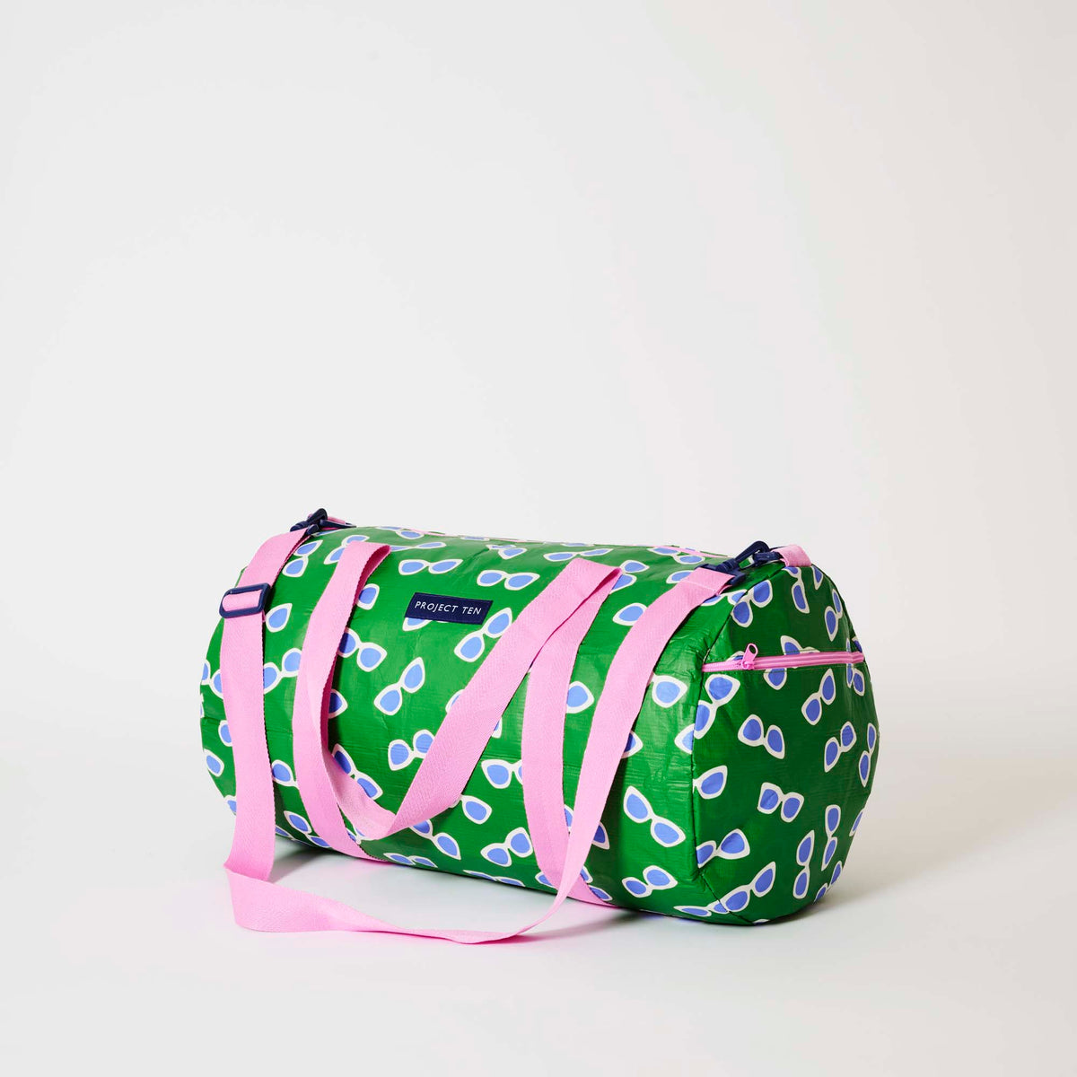 Our Sunglasses Barrel Bag, a cylindrical duffle-style bag in green with a scattered print of blue-lensed, white-framed sunglasses, two pink carry handles, a detachable pink shoulder strap and a pink zip.
