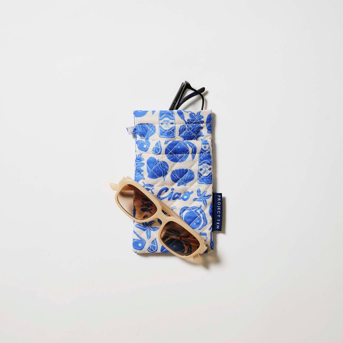 Our Feast Double Glasses Case, a soft quilted glasses case in an ecru base with a blue seaside print of shells, sardines, oranges, soda bottles and "Ciao" lettering, with a squeeze-top opening and a small loop tab.