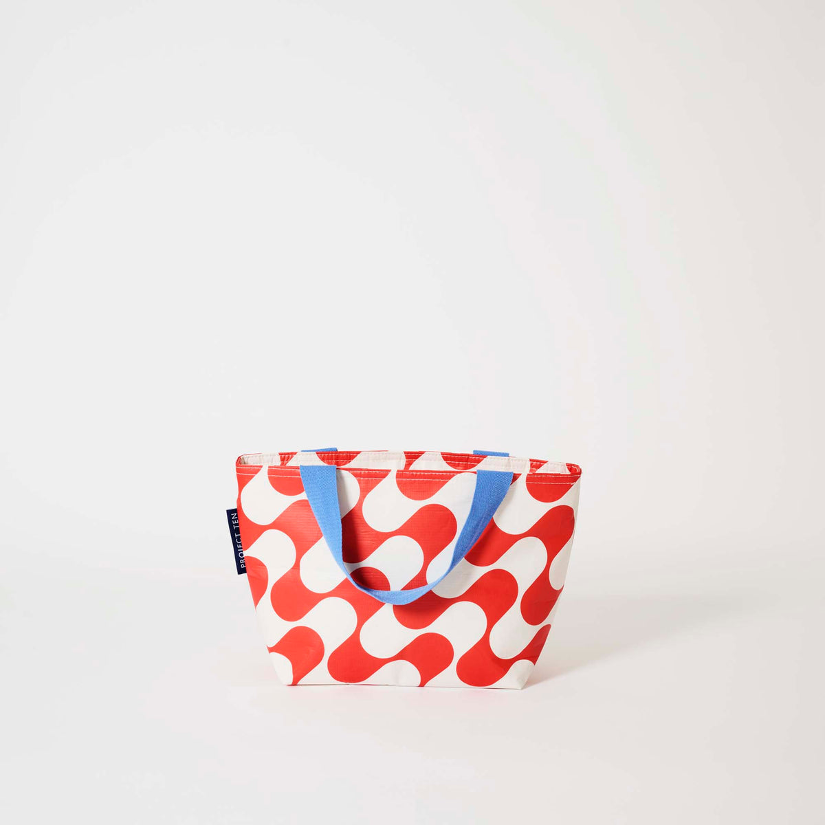 Our Red Ripple Mini Tote, a compact tote bag in a bold geometric wave print of red and ecru, with two blue carry handles.