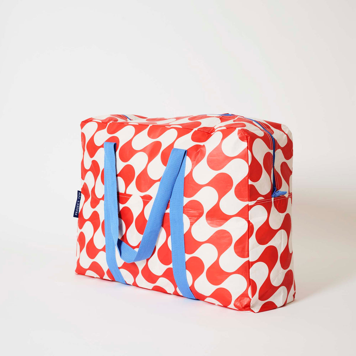 Our Red Ripple Overnight Bag, a roomy duffle-style bag in a bold geometric wave print of red and ecru, with two blue carry handles and a blue zip.