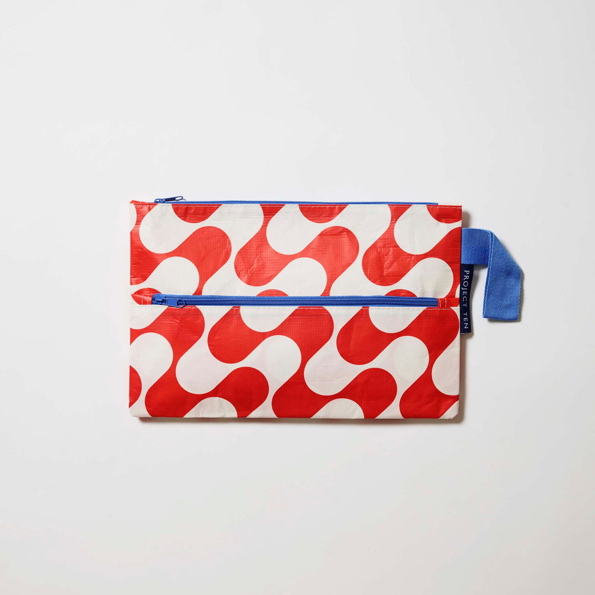 Our Red Ripple Pencil Case, a flat rectangular case in a red and ecru wave print with two blue zip compartments and a blue handle.