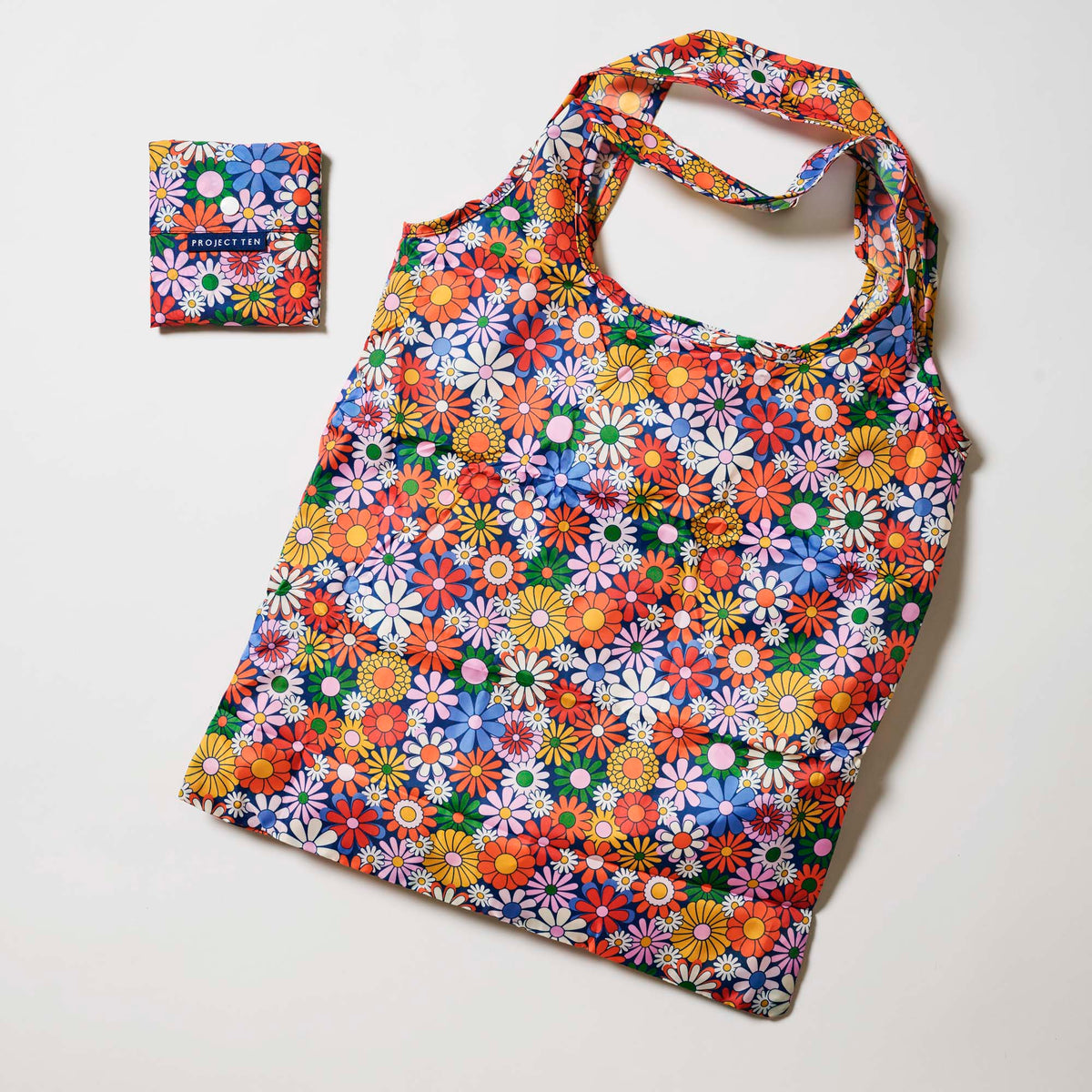 Our Coastal Floral Pocket Shopper, a lightweight foldable tote covered in bright retro daisies in orange, pink, yellow and blue on a navy background, shown open and laid flat next to its matching pouch that it folds into with a snap closure.