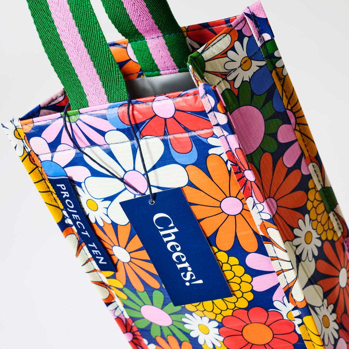 Our Coastal Floral Wine Bag, a tall narrow bottle bag in a retro daisy print of orange, red, pink, yellow and white flowers on a navy background, with a single green and pink striped carry handle.