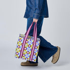 A woman in a blue denim jacket and wide-leg jeans walks in profile while carrying a colorful patterned tote bag with purple handles against a plain light gray studio background.