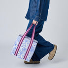 A model in a blue denim outfit carries a large white tote bag with a blue floral pattern and pink handles, shown from a side profile against a light gray studio background.