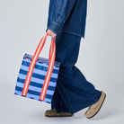 A model in a blue denim outfit walks while carrying a blue and white striped tote bag with red-orange handles against a light gray studio background.