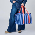 A woman stands against a light gray studio background wearing a blue denim shirt and wide-leg jeans, holding a blue striped tote bag with red handles.