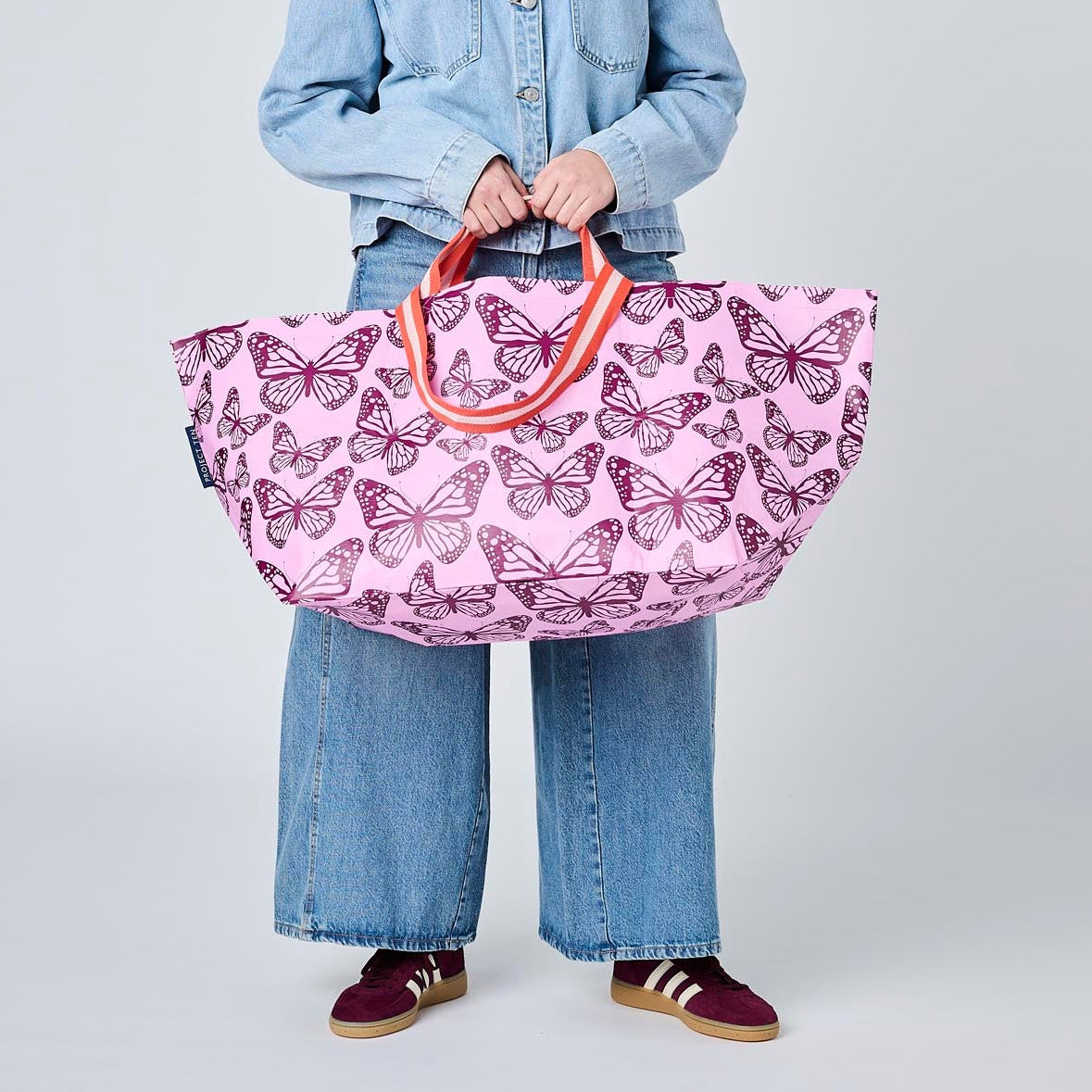 A model wearing a light blue denim jacket and jeans holds a large pink tote bag decorated with a purple butterfly pattern. The background is a clean, neutral gray studio backdrop.