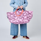 A model wearing a light blue denim jacket and jeans holds a large pink tote bag decorated with a purple butterfly pattern. The background is a clean, neutral gray studio backdrop.