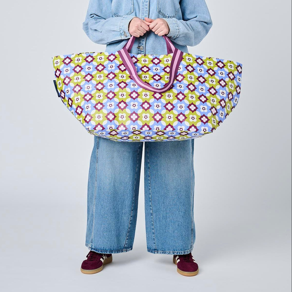 A model wearing a light blue denim shirt and wide-leg jeans holds a large, colorful patterned duffel bag with purple handles against a plain light gray background.