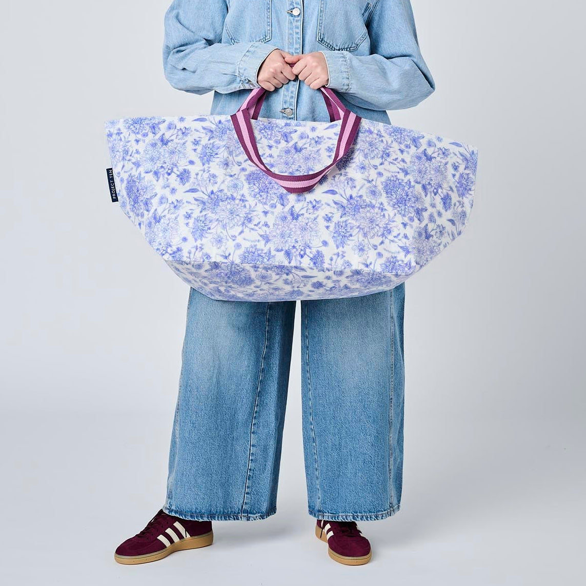 A model wearing a light blue denim shirt and wide-leg jeans holds a large, soft-sided duffel bag with a blue and white floral pattern and purple handles, set against a plain light gray studio background.