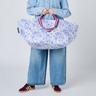 A model wearing a light blue denim shirt and wide-leg jeans holds a large, soft-sided duffel bag with a blue and white floral pattern and purple handles, set against a plain light gray studio background.