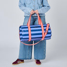 A model stands against a light gray studio background wearing a light blue denim jacket and wide-leg jeans, holding a blue and white striped duffel bag with orange-red handles.