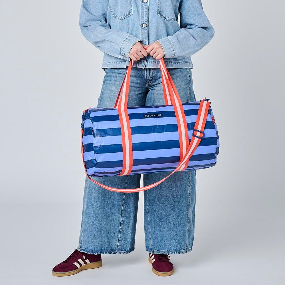 A model stands against a light gray studio background wearing a light blue denim jacket and wide-leg jeans, holding a blue and white striped duffel bag with orange-red handles.