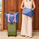 Woman standing with a suitcase and two polka dot bags in front of a wooden building.