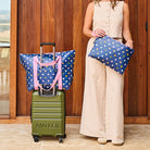 Woman standing with a green suitcase and two polka dot bags in front of a wooden building.
Bag on suitcase is zip medium tote and lady is holding the large zip pouch. 