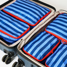 Four blue and navy striped packing cubes with bright red zippers neatly arranged inside an open gray suitcase.