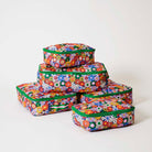 Our Coastal Floral Packing Cubes, a set of five zip-up packing cubes in different sizes, in a retro daisy print of orange, red, pink, yellow, blue and white flowers on a navy background, with green zips and green and pink striped carry handles.