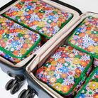 Our Coastal Floral Packing Cubes, a set of five zip-up packing cubes in different sizes, in a retro daisy print of orange, red, pink, yellow, blue and white flowers on a navy background, with green zips and green and pink striped carry handles.