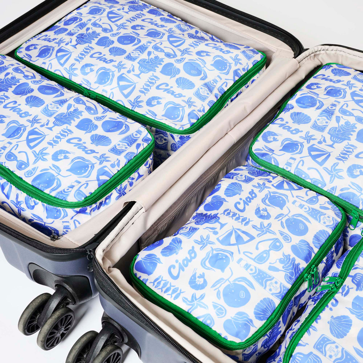 Our Feast Packing Cubes, a set of five zip-up packing cubes in different sizes, in an ecru base with a blue seaside print of shells, oranges, soda bottles, sunglasses and "Ciao" lettering, with green zips and green and pink striped carry handles.