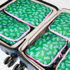 Our Sunglasses Packing Cubes, a set of five zip-up packing cubes in different sizes, in green with a scattered print of blue-lensed, white-framed sunglasses, with pink zips and pink carry handles.