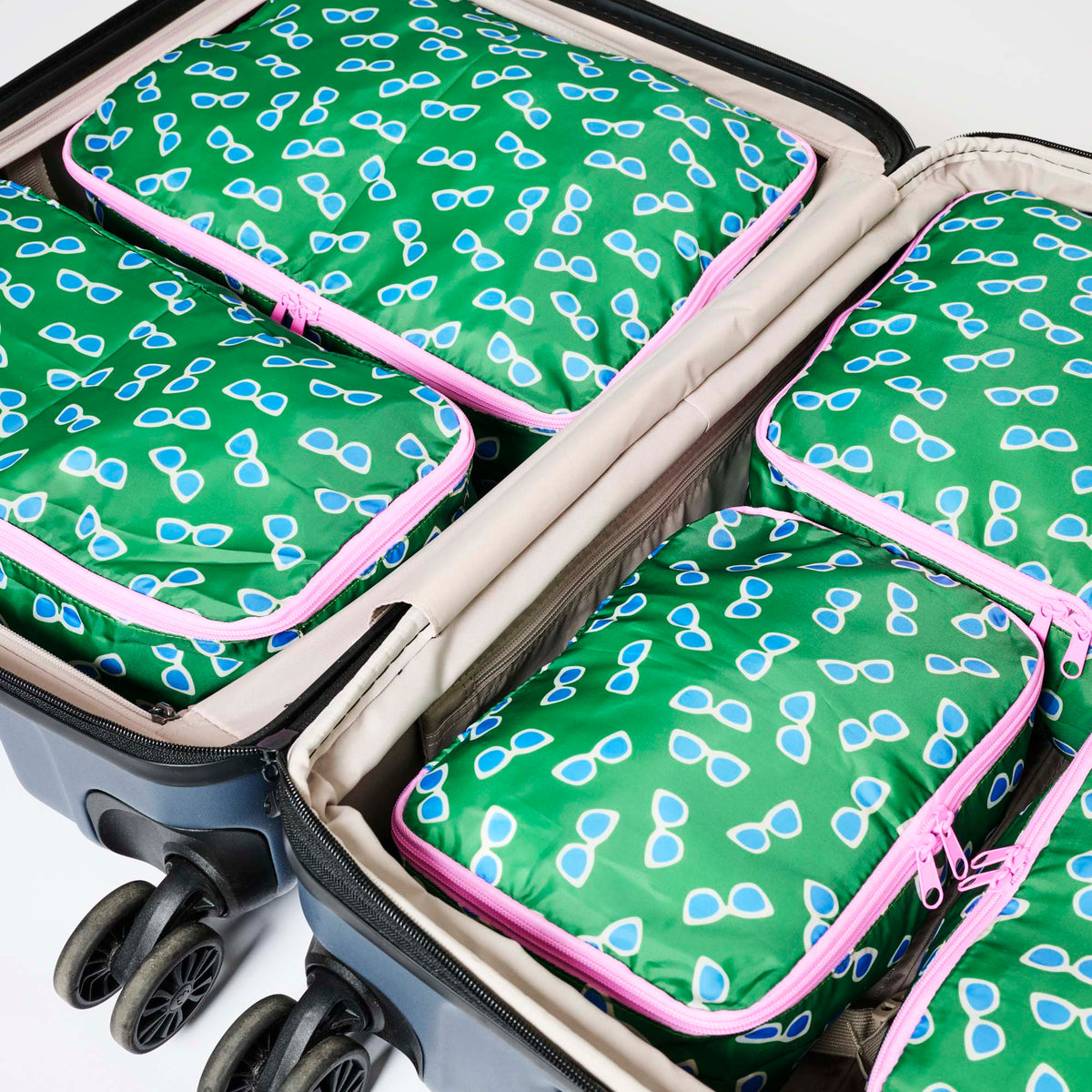 Our Sunglasses Packing Cubes, a set of five zip-up packing cubes in different sizes, in green with a scattered print of blue-lensed, white-framed sunglasses, with pink zips and pink carry handles.