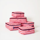 A set of five packing cubes stacked in a pyramid arrangement, featuring a vibrant pink, red, and white striped pattern with small dotted details and dark purple trim.
- Project Ten