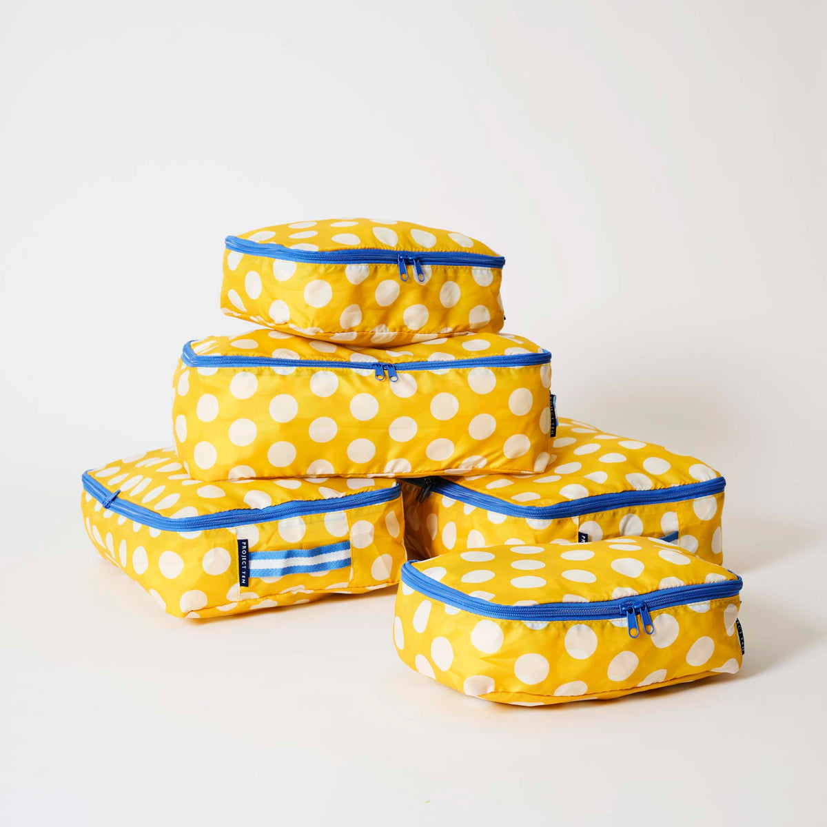 Our Yellow Polka Dot Packing Cubes, a set of five zip-up packing cubes in different sizes, in bright yellow with large ecru polka dots, with blue zips and blue and ecru striped carry handles.