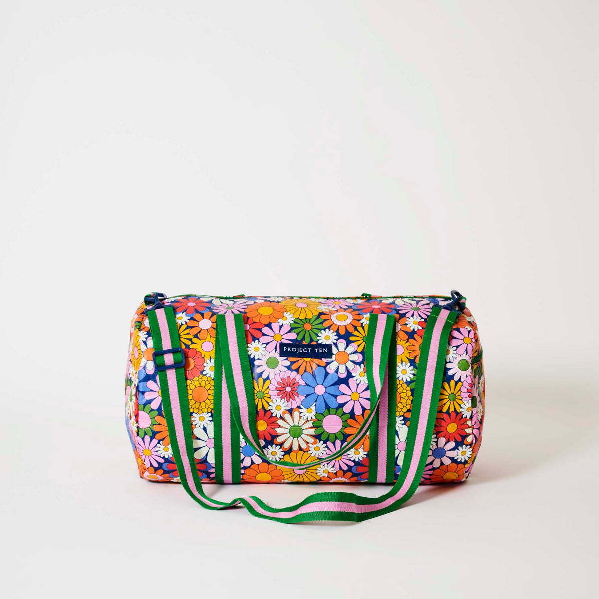 Our Coastal Floral Barrel Bag, a cylindrical duffle-style bag in a retro daisy print of orange, red, pink, yellow, blue and white flowers on a navy background, with two green and pink striped carry handles, a detachable matching shoulder strap and a green zip.
