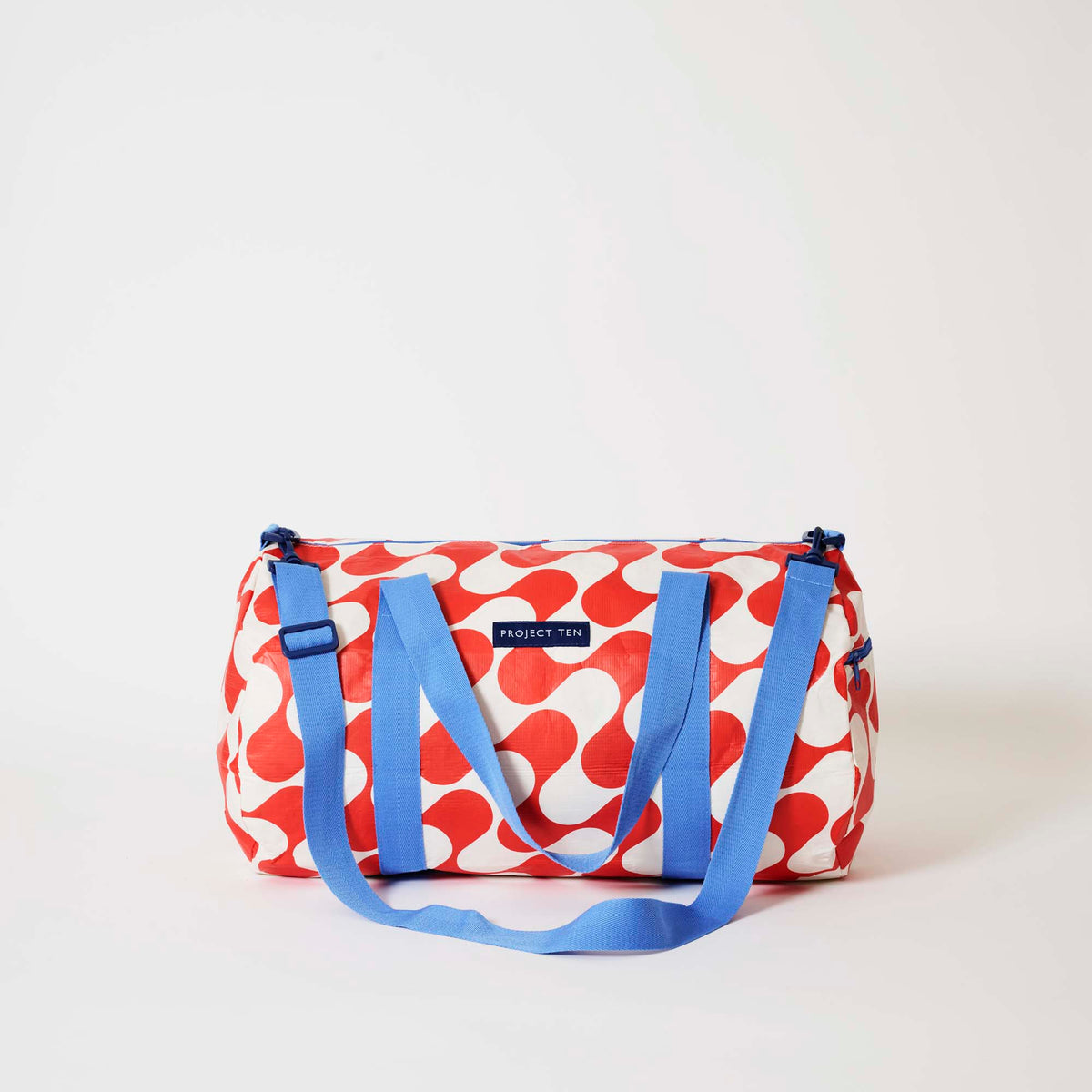 Our Red Ripple Barrel Bag, a cylindrical duffle-style bag in a bold geometric wave print of red and ecru, with two blue carry handles, a detachable blue shoulder strap and a blue zip.