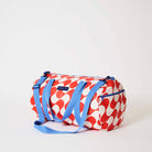 Our Red Ripple Barrel Bag, a cylindrical duffle-style bag in a bold geometric wave print of red and ecru, with two blue carry handles, a detachable blue shoulder strap and a blue zip.