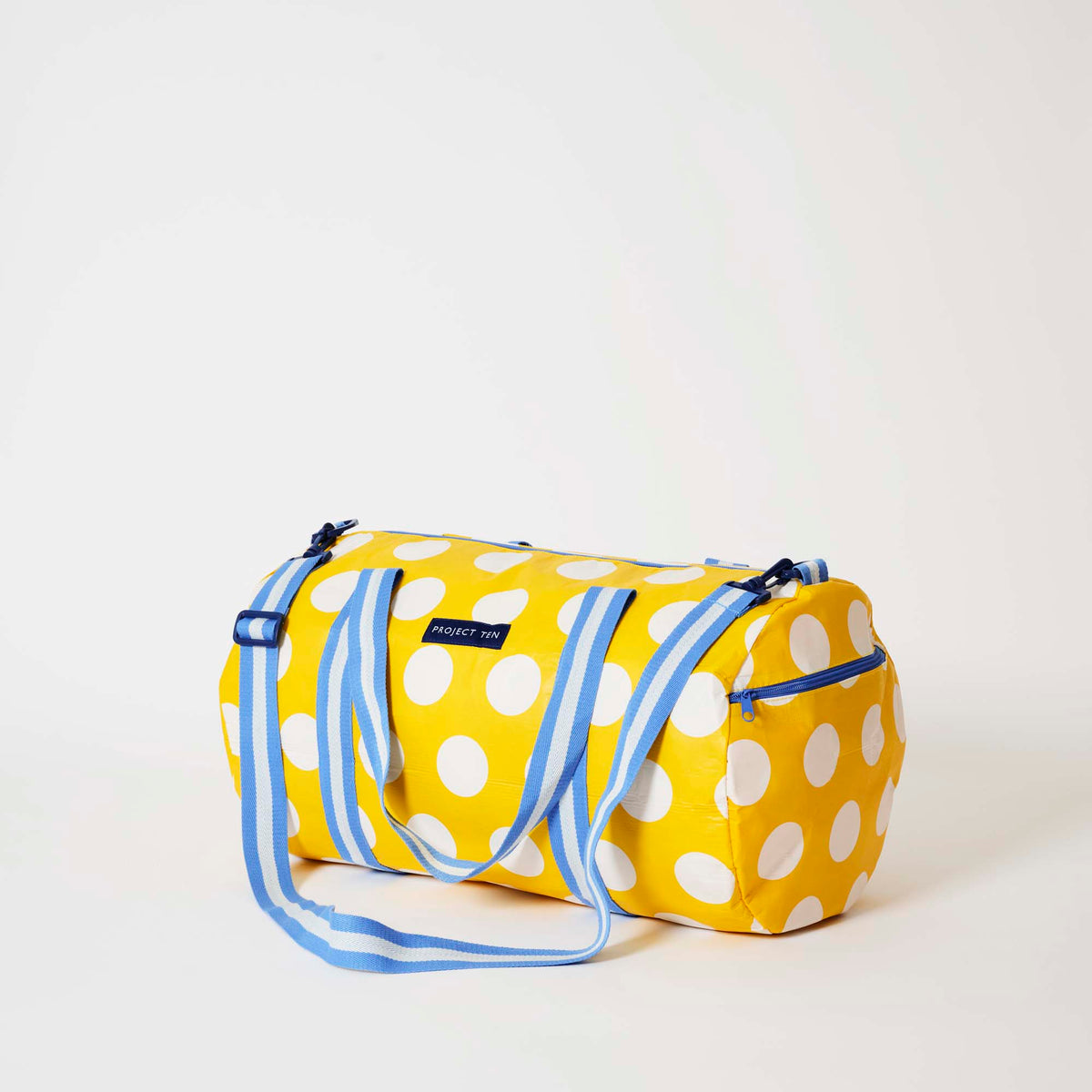 Our Yellow Polka Dot Barrel Bag, a cylindrical duffle-style bag in bright yellow with large ecru polka dots, two blue and ecru striped carry handles, a detachable matching shoulder strap and a blue zip.
