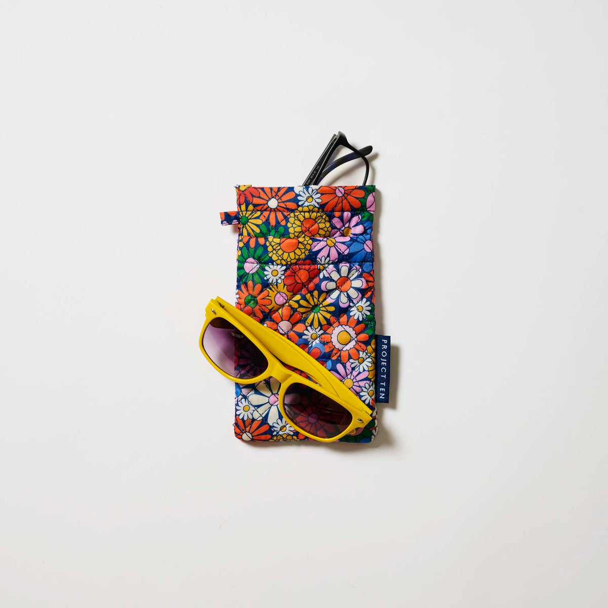 Our Coastal Floral Double Glasses Case, a soft quilted glasses case in a retro daisy print of orange, red, pink, yellow, blue and white flowers on a navy background, with a squeeze-top opening and a small loop tab.