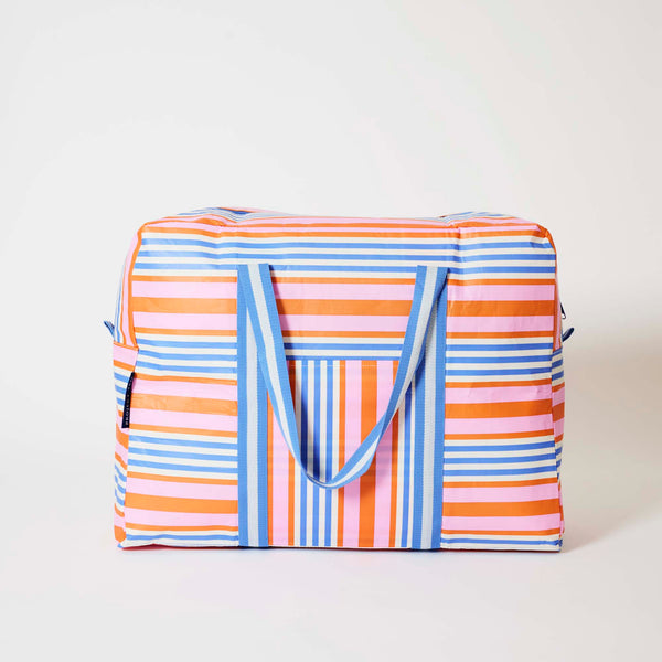 Our Capri Stripe Overnight Bag, a roomy duffle-style bag in pink, orange, blue and ecru stripes, with a vertical striped front pocket, two blue and ecru striped carry handles and a blue zip.