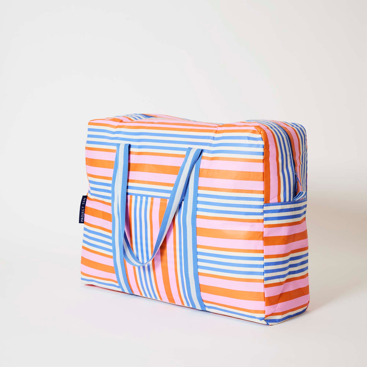 Our Capri Stripe Overnight Bag, a roomy duffle-style bag in pink, orange, blue and ecru stripes, with a vertical striped front pocket, two blue and ecru striped carry handles and a blue zip.