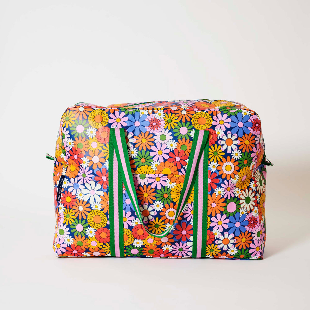 Our Coastal Floral Overnight Bag, a roomy duffle-style bag in a retro daisy print of orange, red, pink, yellow, blue and white flowers on a navy background, with two green and pink striped carry handles and a green zip.